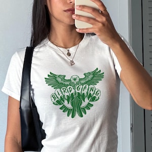 Może przedstawiać: Biały t-shirt z zieloną grafiką orła i napisem "BIRD GANG". Koszulka ma kr&oacute;tkie rękawy i okrągły dekolt. Projekt znajduje się z przodu koszulki.