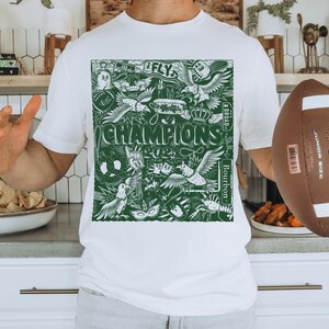 May include: White t-shirt featuring a green graphic print of a football team's logo. The logo includes the words "Fly", "Champions", "Bourbon", and "4 & Broad".