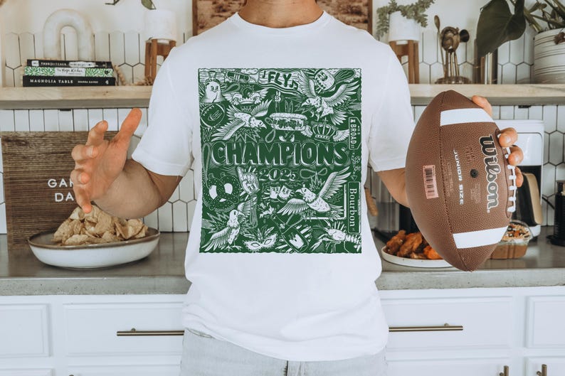 May include: White t-shirt featuring a green graphic print of a football team's logo. The logo includes the words "Fly", "Champions", "Bourbon", and "4 & Broad".
