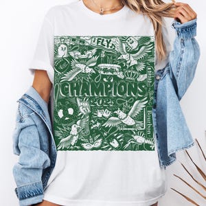 May include: White t-shirt featuring a green graphic print of various images, including a pretzel, a crown, a bird, and the text "FLY" and "CHAMPIONS 2023".