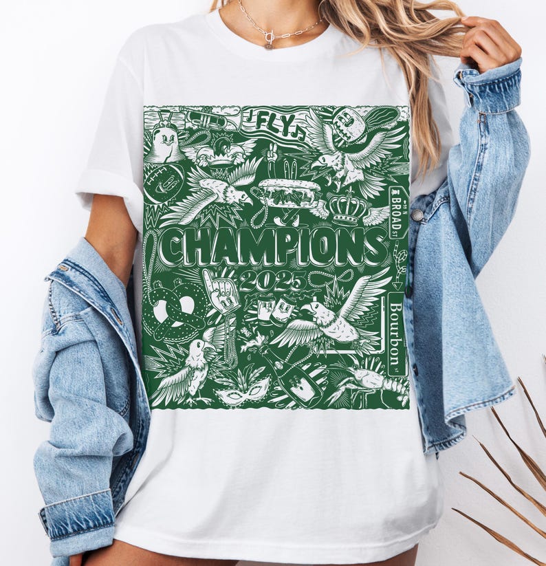 May include: White t-shirt featuring a green graphic print of various images, including a pretzel, a crown, a bird, and the text "FLY" and "CHAMPIONS 2023".