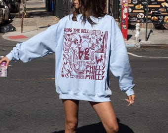 May include: A light blue sweatshirt with a graphic print of baseball-themed illustrations and the text "Ring the Bell" and "Philly" repeated three times.