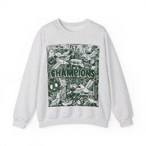 Puede incluir: Una sudadera gris con un estampado gr&aacute;fico verde de varias im&aacute;genes, incluyendo p&aacute;jaros, una corona y el texto "CHAMPIONS 2025".