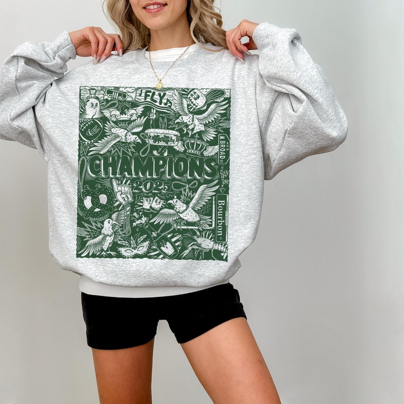 Puede incluir: Una sudadera gris con un estampado gr&aacute;fico verde de una variedad de im&aacute;genes, incluyendo un p&aacute;jaro, una corona, una m&aacute;scara y las palabras "FLY" y "CHAMPIONS 2023".