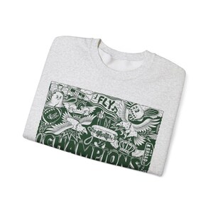Puede incluir: Una sudadera gris con un estampado gr&aacute;fico verde de un &aacute;guila de dibujos animados, una corona y las palabras "Fly" y "Champions". El gr&aacute;fico tambi&eacute;n incluye el texto "Broad St."