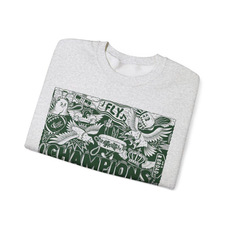 Puede incluir: Una sudadera gris con un estampado gr&aacute;fico verde de un &aacute;guila de dibujos animados, una corona y las palabras "Fly" y "Champions". El gr&aacute;fico tambi&eacute;n incluye el texto "Broad St."