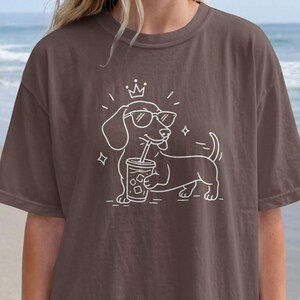 May include: Espresso-colored t-shirt featuring a white line drawing of a dachshund wearing sunglasses and a crown, holding a beverage. The shirt is a relaxed fit. The word "ESPRESSO" is in a brown circle on the bottom right.