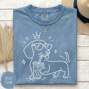 May include: Blue jean colored t-shirt with a white graphic of a dachshund wearing sunglasses and a crown, holding a drink with a straw. The shirt has a Comfort Colors tag and a silver necklace.