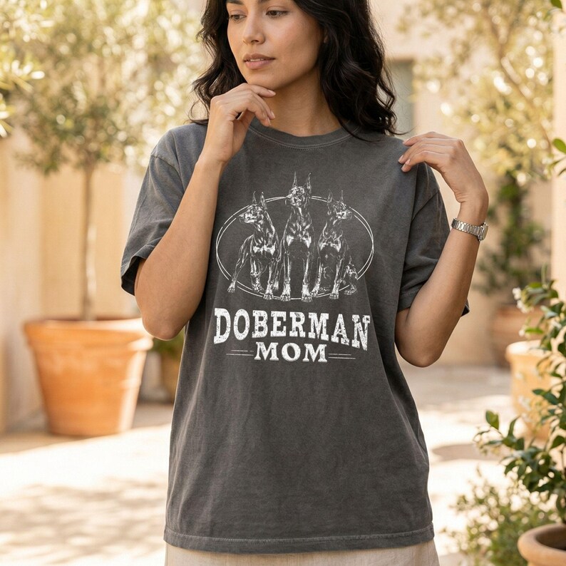 May include: Charcoal grey t-shirt with a white graphic of three Doberman Pinschers in an oval, and the words "DOBERMAN MOM". The shirt has a relaxed fit and short sleeves.