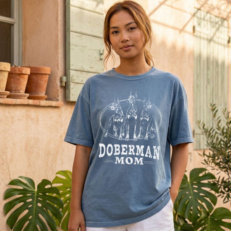 May include: A blue t-shirt with a white graphic of three Doberman Pinschers inside an oval, with the words "DOBERMAN MOM" below. The shirt is a solid color and has short sleeves.