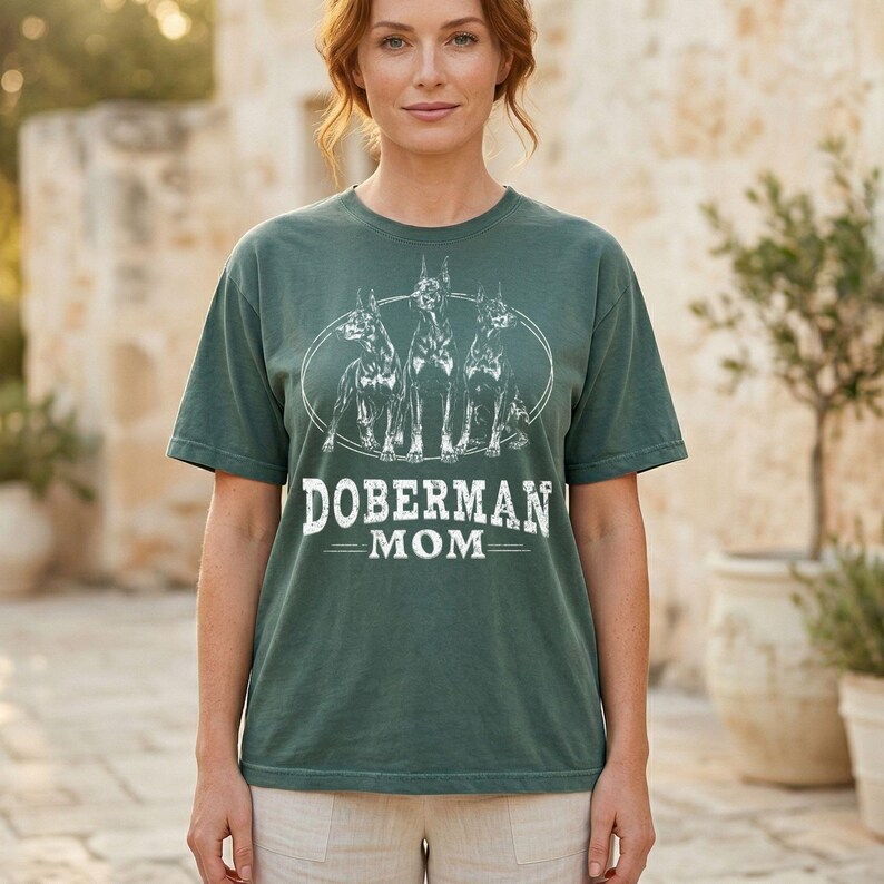May include: A green t-shirt with a white graphic of three Doberman Pinschers in an oval, and the text "DOBERMAN MOM" underneath. The shirt is a solid green colour and has short sleeves.