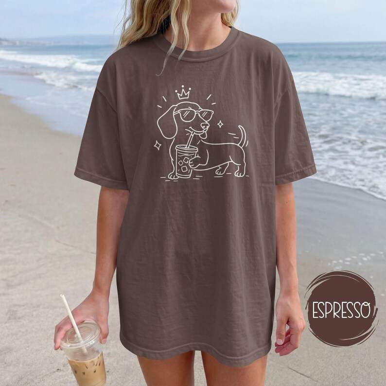 May include: Espresso-colored t-shirt featuring a white line drawing of a dachshund wearing sunglasses and a crown, holding a beverage. The shirt is a relaxed fit. The word "ESPRESSO" is in a brown circle on the bottom right.