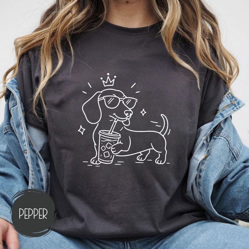May include: Dark grey t-shirt with a white outline of a dachshund wearing sunglasses and a crown, holding a drink with a straw. The word "Pepper" is printed on a dark grey circle at the bottom left.