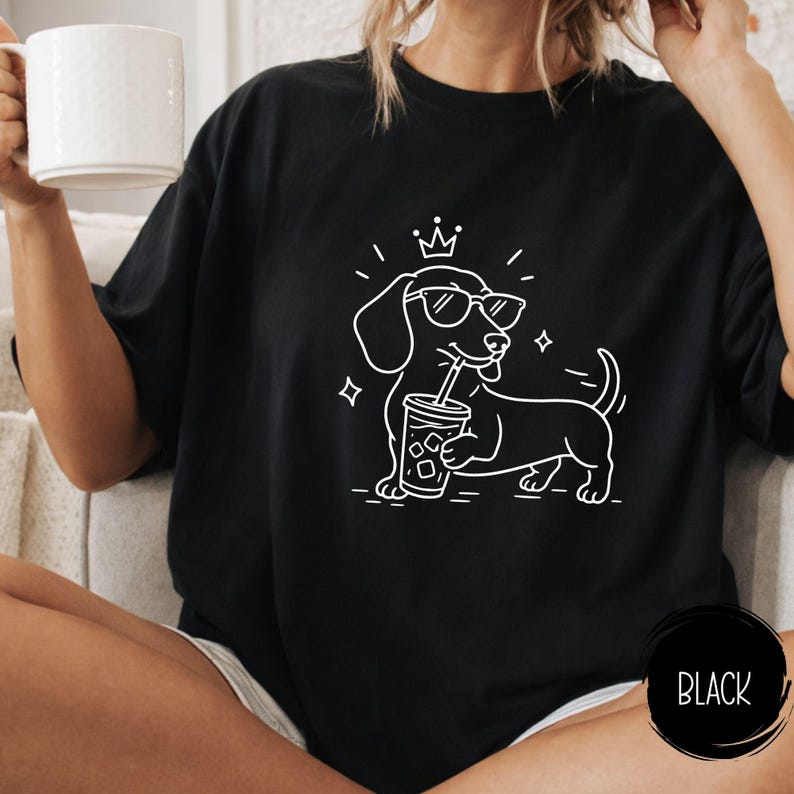 May include: Black t-shirt featuring a white outline illustration of a dachshund wearing sunglasses and a crown, holding a drink with a straw. The word "BLACK" is in a black circle at the bottom right.