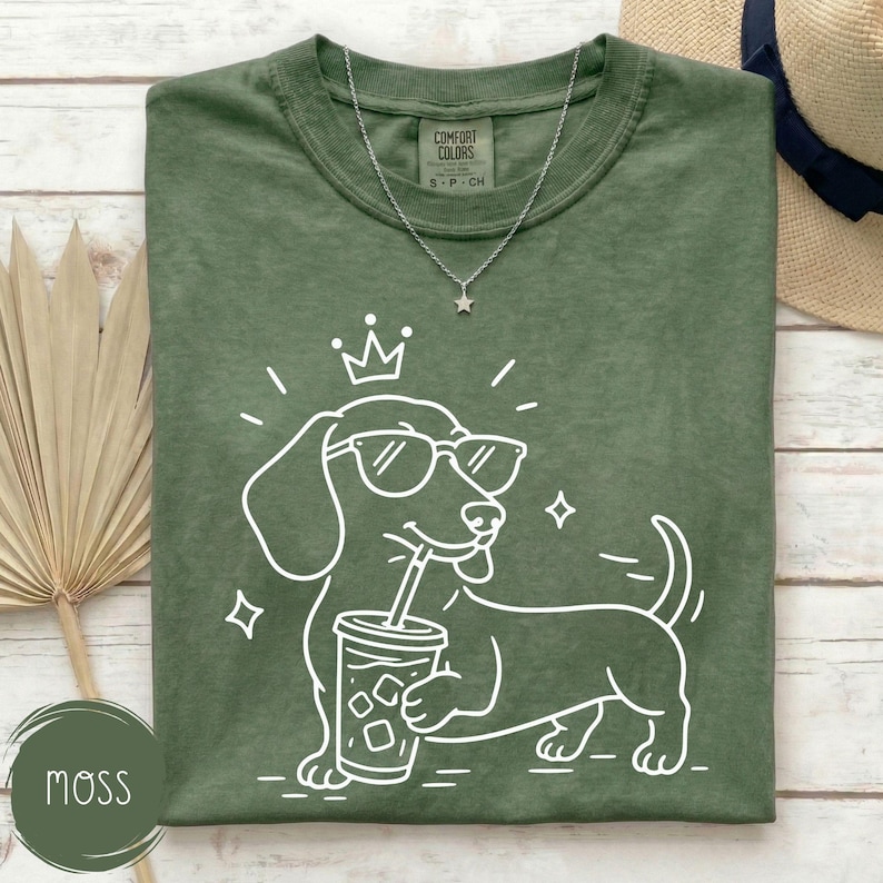 May include: A moss green t-shirt with a white graphic of a dachshund wearing sunglasses and a crown, holding a drink with a straw. A silver necklace with a star pendant is on the shirt. The shirt has the text "Comfort Colors" on the tag.