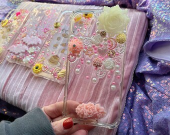 May include: A clear phone case decorated with pink, white, and yellow flowers, pearls, and glitter. The case is on a pink and white striped fabric.