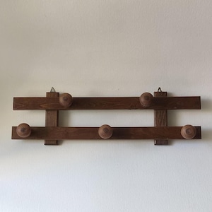 Puede incluir: Un perchero de madera marrón oscuro con dos barras horizontales y cinco pomos de madera. El perchero está montado en una pared blanca y es ideal para colgar abrigos.
