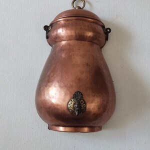 May include: A copper water dispenser with a decorative spout and a lid with a ring handle. The dispenser is hanging on a white wall.