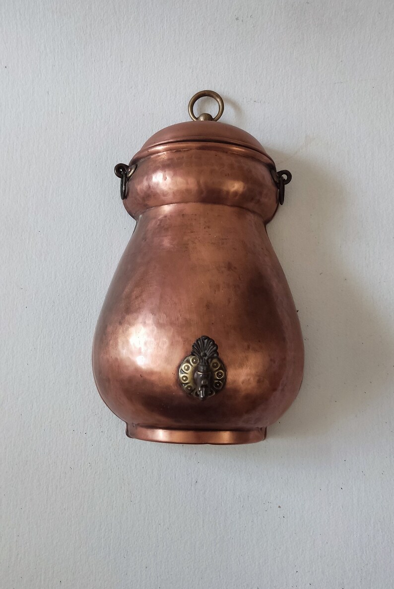 May include: A copper water dispenser with a decorative spout and a lid with a ring handle. The dispenser is hanging on a white wall.
