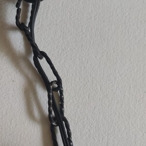 May include: Close-up of a dark metal chain with a decorative element. The chain is made of twisted links and connects to a larger, possibly decorative, object. The background is a plain, off-white surface.
