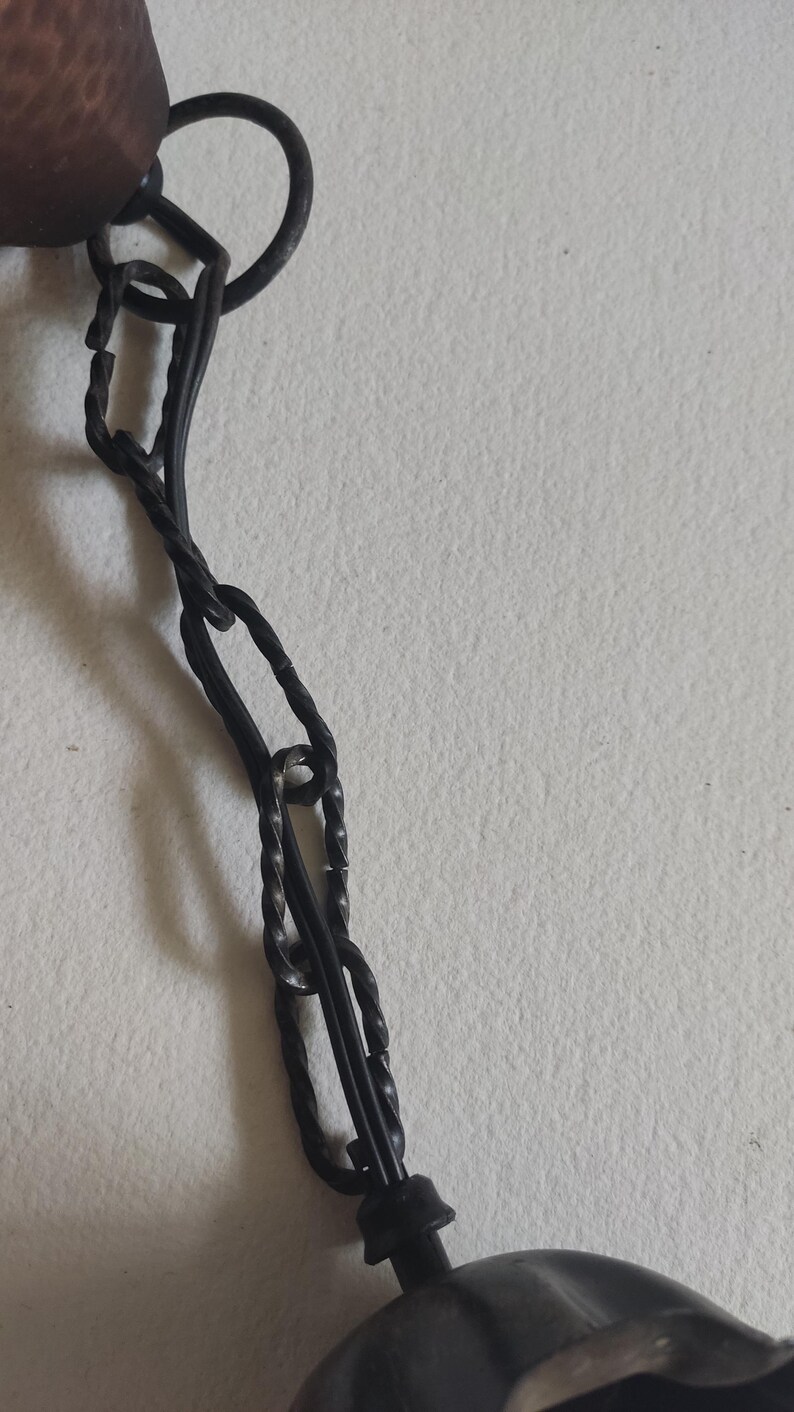 May include: Close-up of a dark metal chain with a decorative element. The chain is made of twisted links and connects to a larger, possibly decorative, object. The background is a plain, off-white surface.