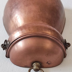 May include: A copper wall-mounted pot with a hinged lid and a decorative handle. The pot has a rounded shape and a smooth, polished surface.