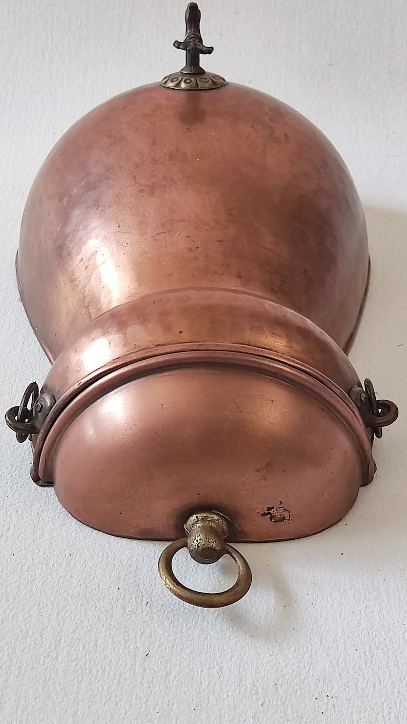 May include: A copper wall-mounted pot with a hinged lid and a decorative handle. The pot has a rounded shape and a smooth, polished surface.