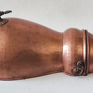 May include: A copper and brass antique water pitcher with a hinged lid and a spout on top. The pitcher has a hammered finish and a ring handle.