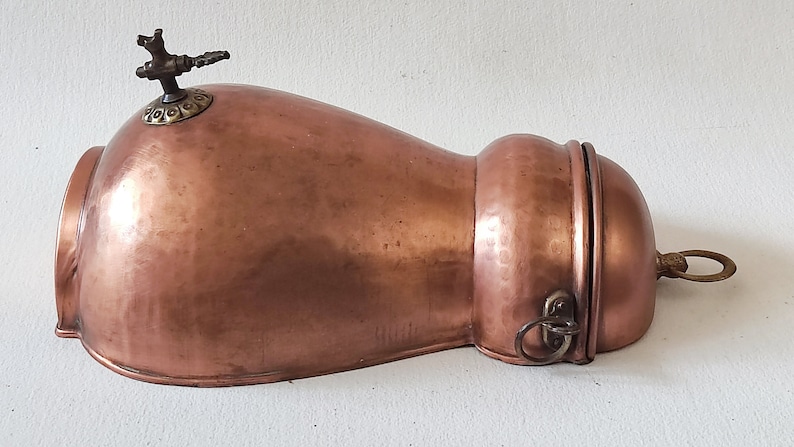 May include: A copper and brass antique water pitcher with a hinged lid and a spout on top. The pitcher has a hammered finish and a ring handle.