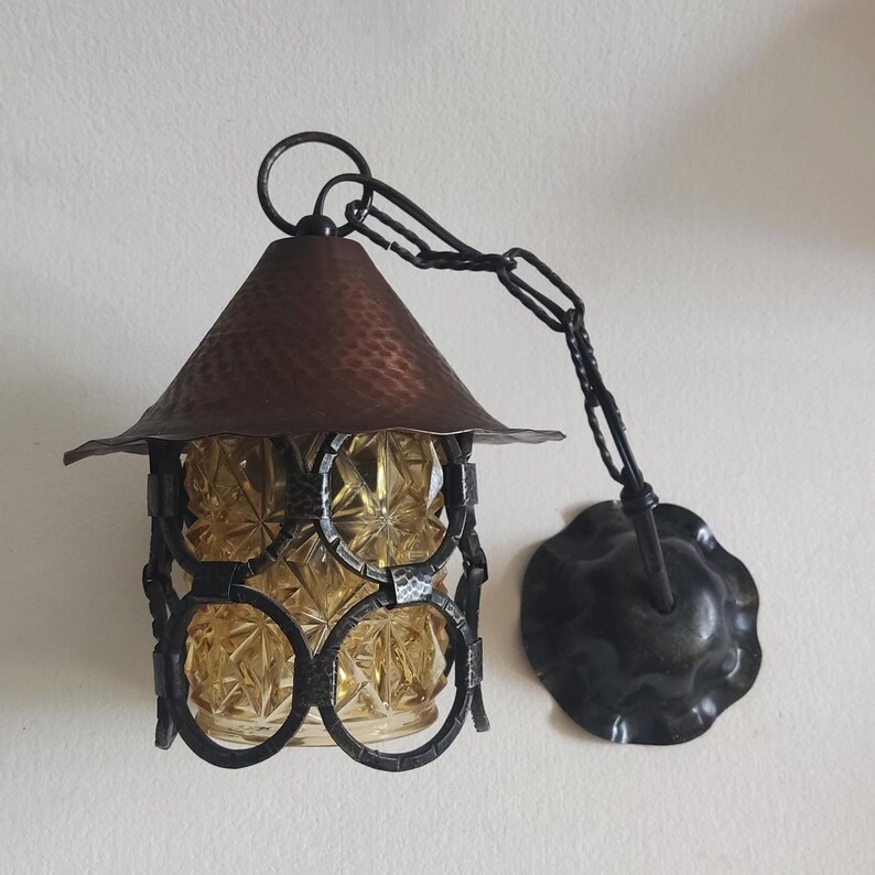 May include: An antique hanging lantern with a copper-colored, textured roof and a decorative, dark metal frame. The lantern features a chain for hanging and a circular wall mount. The glass inside the lantern is amber-colored.