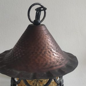 May include: Close-up of a decorative hanging lantern with a copper-colored, textured shade and a dark metal frame. The lantern has a ring at the top for hanging. The bottom portion of the lantern is visible, with a yellow-tinted glass design.