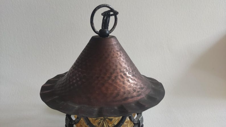 May include: Close-up of a decorative hanging lantern with a copper-colored, textured shade and a dark metal frame. The lantern has a ring at the top for hanging. The bottom portion of the lantern is visible, with a yellow-tinted glass design.