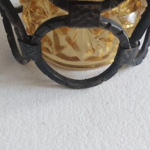 May include: Close-up of a vintage amber glass bowl with a starburst pattern, encased in a dark metal filigree frame. The bowl has a textured surface and is partially visible, showcasing its intricate design. The metal frame adds a decorative element.