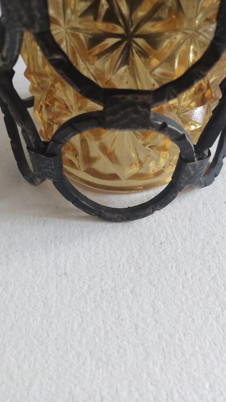 May include: Close-up of a vintage amber glass bowl with a starburst pattern, encased in a dark metal filigree frame. The bowl has a textured surface and is partially visible, showcasing its intricate design. The metal frame adds a decorative element.