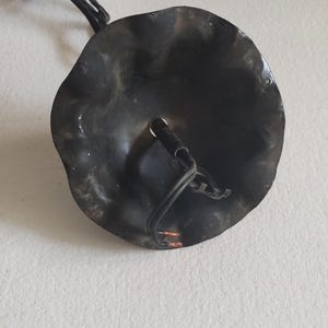 May include: A black, metal ceiling light fixture with a wavy, circular shade. The fixture has a chain for hanging and a wire for electrical connection. The shade has a textured surface and a hole for the light bulb.