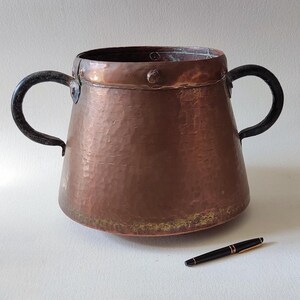 Puede incluir: Una olla grande de cobre martillado con dos asas de hierro negro. La olla tiene un fondo redondeado y una abertura ancha y acampanada.