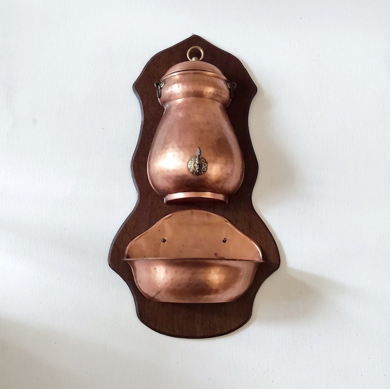 May include: A copper wall-mounted water fountain with a wooden frame. The fountain has a copper pitcher with a lid and a spout, and a copper basin below.