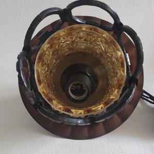 May include: Overhead view of a vintage light fixture. The fixture features a brown, circular base with a decorative, amber-colored glass shade. A dark metal frame with a chain-like design surrounds the shade. The light socket is visible inside the shade.