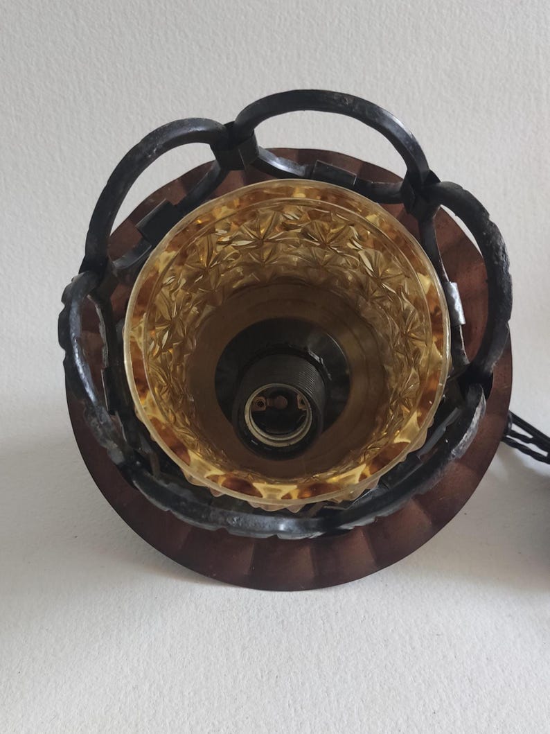 May include: Overhead view of a vintage light fixture. The fixture features a brown, circular base with a decorative, amber-colored glass shade. A dark metal frame with a chain-like design surrounds the shade. The light socket is visible inside the shade.