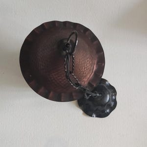 May include: A copper-colored, hammered metal pendant light fixture. The fixture has a circular shade with a scalloped edge, a chain, and a decorative ceiling plate. The metal has a textured surface and a dark, aged finish.