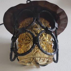 May include: An antique hanging lamp with a decorative amber glass shade and a dark metal frame. The shade has a diamond pattern, and the frame features circular and curved elements. The lamp is designed to be suspended from a ceiling.