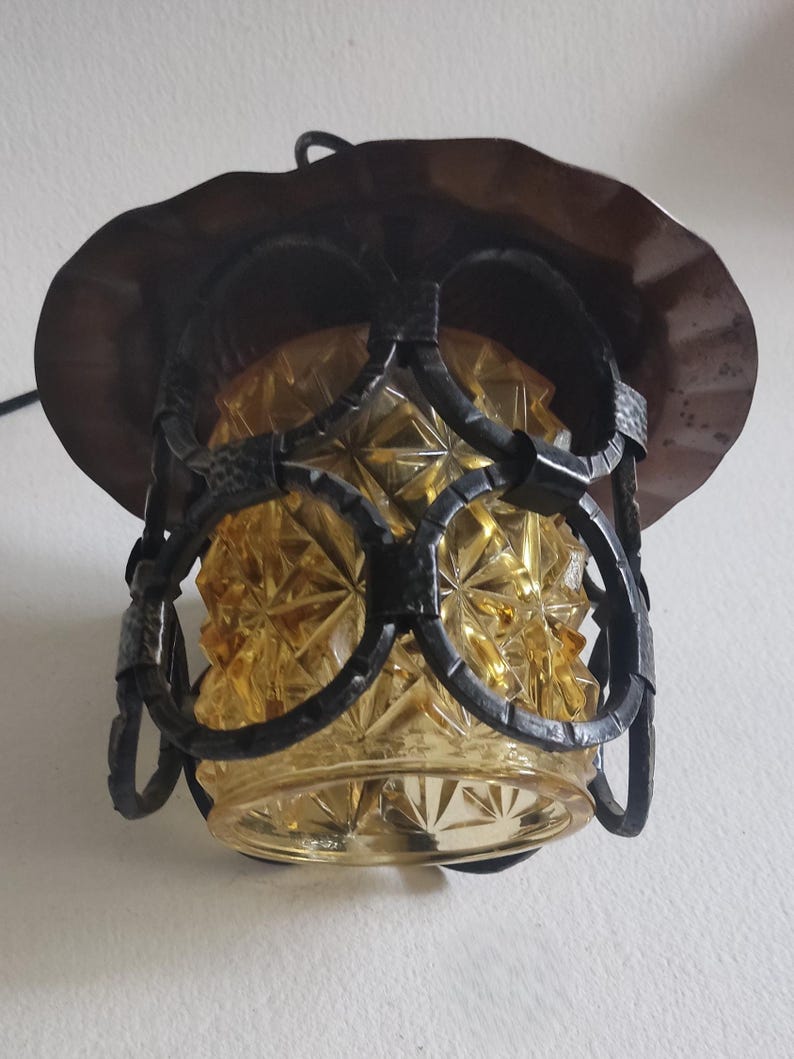 May include: An antique hanging lamp with a decorative amber glass shade and a dark metal frame. The shade has a diamond pattern, and the frame features circular and curved elements. The lamp is designed to be suspended from a ceiling.