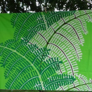 May include: A vibrant green fabric featuring a large fern leaf design in green, white, and brown. The fabric is displayed outdoors, highlighting the striking botanical pattern. This fabric is suitable for home decor or craft projects.