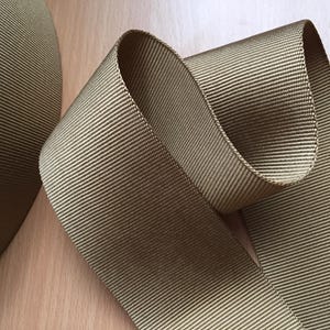 May include: A close-up of a roll of brown webbing. The webbing is woven with a textured pattern and has a smooth, matte finish.