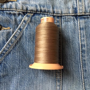 May include: A spool of brown thread on a blue denim background. The thread is labelled "Gütermann 100% Polyester". The spool has a light peach-coloured base and top. The denim jacket has visible stitching and metal buttons.