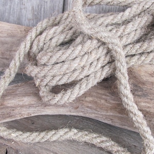 Könnte beinhalten: Aufgerolltes Naturfaserseil, wahrscheinlich Hanf oder Jute, mit einer dicken, gedrehten Konstruktion. Das Seil hat eine neutrale beige Farbe und scheint neu zu sein. Der Hintergrund ist eine verwitterte, hellbraune Holzoberfläche.