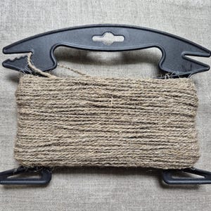 May include: A spool of natural tan jute twine wrapped around a black plastic holder. The holder features a curved handle at the top with notched ends and two smaller, angled feet at the bottom, providing a structured frame for the cord.