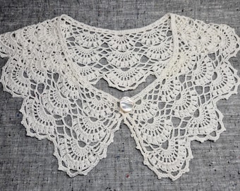May include: Ivory-colored, crocheted collar with a scalloped edge and a button closure. The delicate lace design features a repeating fan pattern. This handmade accessory adds a vintage touch to any outfit.