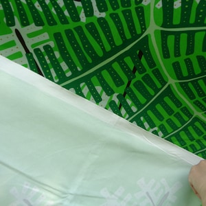 May include: A close-up of a green and white fabric with a leaf pattern. The fabric is a vibrant green with white accents and a repeating leaf design. A white fabric is folded over the green fabric.