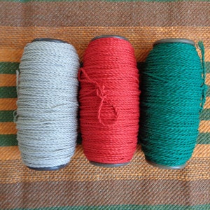 May include: Three spools of rope in gray, red, and green, arranged side by side. The rope is tightly wound around a dark-colored core. The background is a striped fabric in shades of brown, orange, and green.